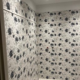 Dariusz Kulig usługi Remontowo-Budowlane - Wykończona łazienka z kwiatowymi płytkami w odcieniach szarości. Widoczny odpływ liniowy w podłodze i narzędzia glazurnicze. Minimalistyczne oświetlenie sufitowe.