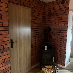 Dariusz Kulig usługi Remontowo-Budowlane - Ściana z cegły klinkierowej w Krakowie, z widocznym piecem wolnostojącym i drewnianymi drzwiami. Detal wykończenia wnętrza z narzędziami i materiałami budowlanymi.
