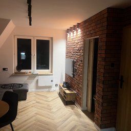 Dariusz Kulig usługi Remontowo-Budowlane - Salon po remoncie: parkiet w jodełkę, ściana z cegły, oświetlenie punktowe. Widok na okno i meble. Nowoczesny design z elementami rustykalnymi.