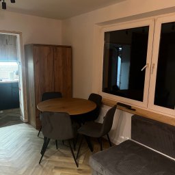 Dariusz Kulig usługi Remontowo-Budowlane - Jasne wnętrze: drewniany stół z krzesłami, parkiet w jodełkę, szafa z forniru i okno z drewnianym parapetem. Widok na kuchnię w tle.