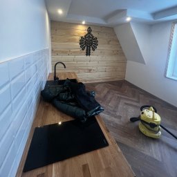 Dariusz Kulig usługi Remontowo-Budowlane - Nowoczesna kuchnia po remoncie: białe płytki, drewniany blat, czarna płyta indukcyjna i zlew. Ściana wykończona drewnem z ozdobnym elementem. Widoczny odkurzacz.