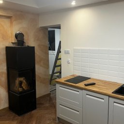 Dariusz Kulig usługi Remontowo-Budowlane - Nowoczesna kuchnia z drewnianym blatem, białymi płytkami i czarnym piecem kominkowym. Parkiet ułożony w jodełkę. W tle drabina.