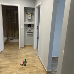 Dariusz Kulig usługi Remontowo-Budowlane - Jasne, drewnopodobne płytki podłogowe w odnowionym wnętrzu. Widoczna kabina prysznicowa, białe futryny drzwi i narzędzia na podłodze. Świeżo pomalowane ściany.