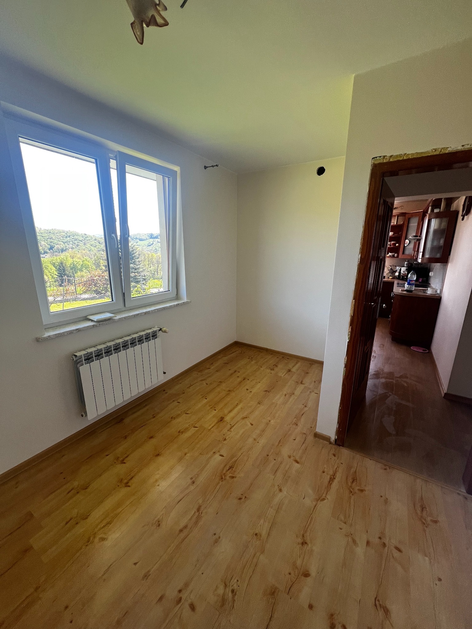 Jasny pokój po malowaniu i położeniu gładzi: drewniana podłoga, białe ściany, okno z widokiem na zieleń, grzejnik. Widoczne przejście do kuchni.