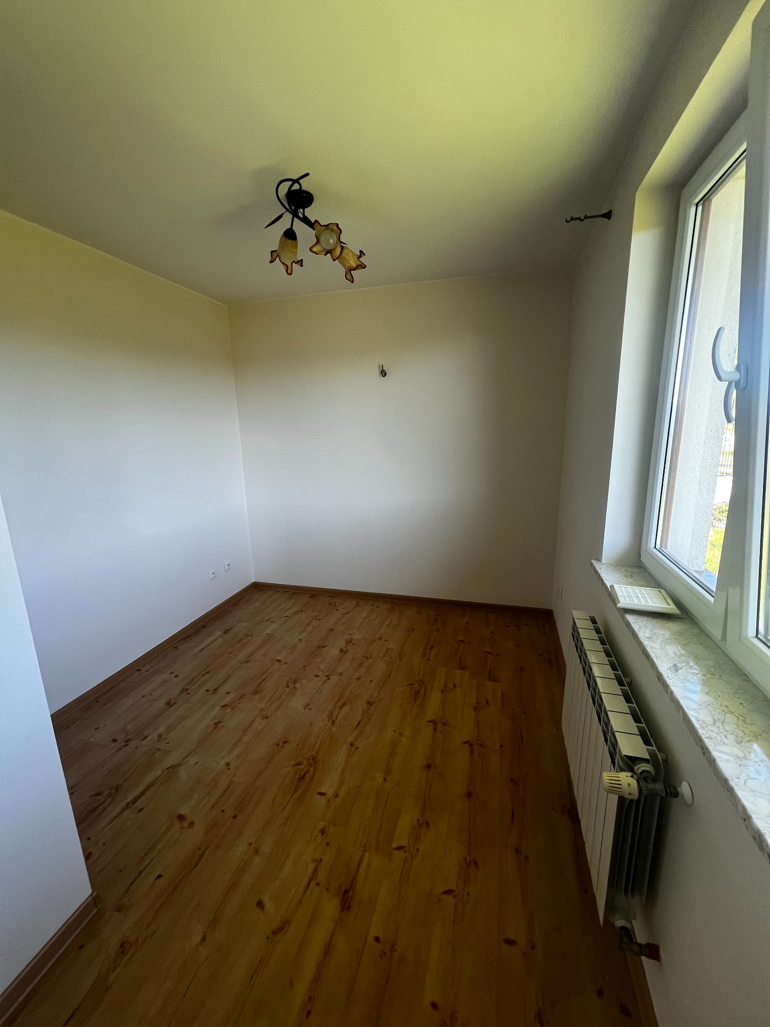 Jasne wnętrze po malowaniu: drewniana podłoga, białe ściany z grzejnikiem pod oknem. Widoczna lampa sufitowa. Świeżo wykończone pomieszczenie.