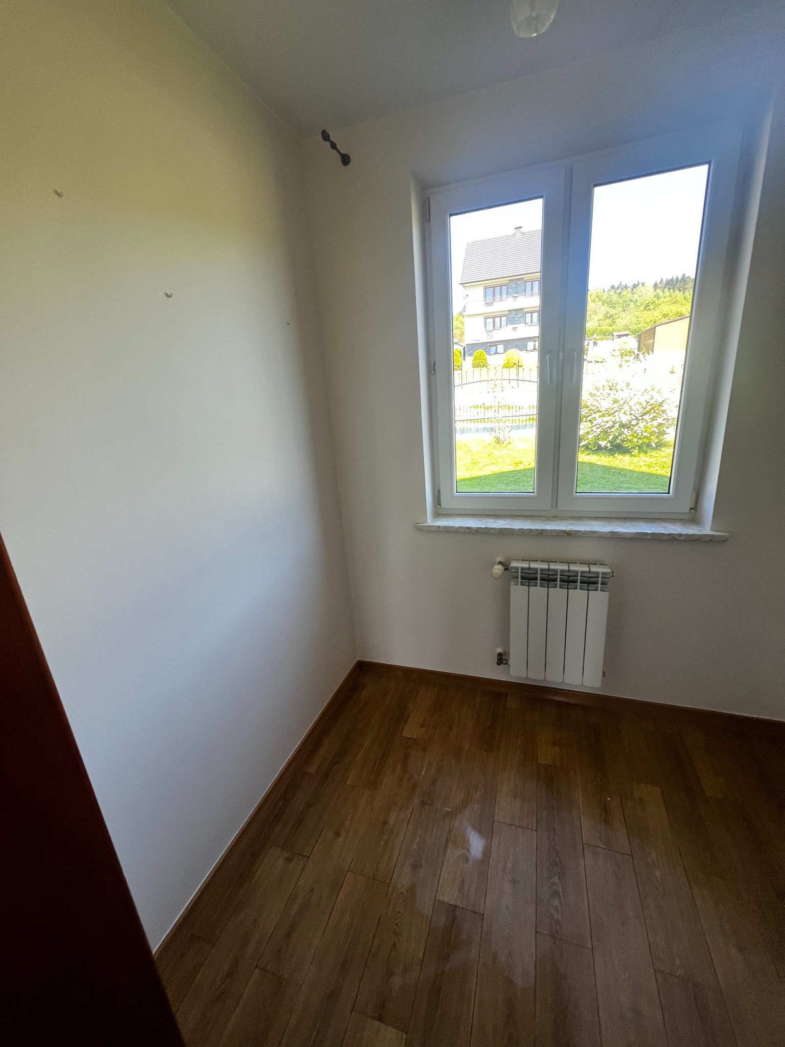Pomalowane na biało wnętrze pokoju z drewnianą podłogą. Widoczne okno z widokiem na dom i zieleń, biały grzejnik pod oknem i fragment brązowych drzwi.
