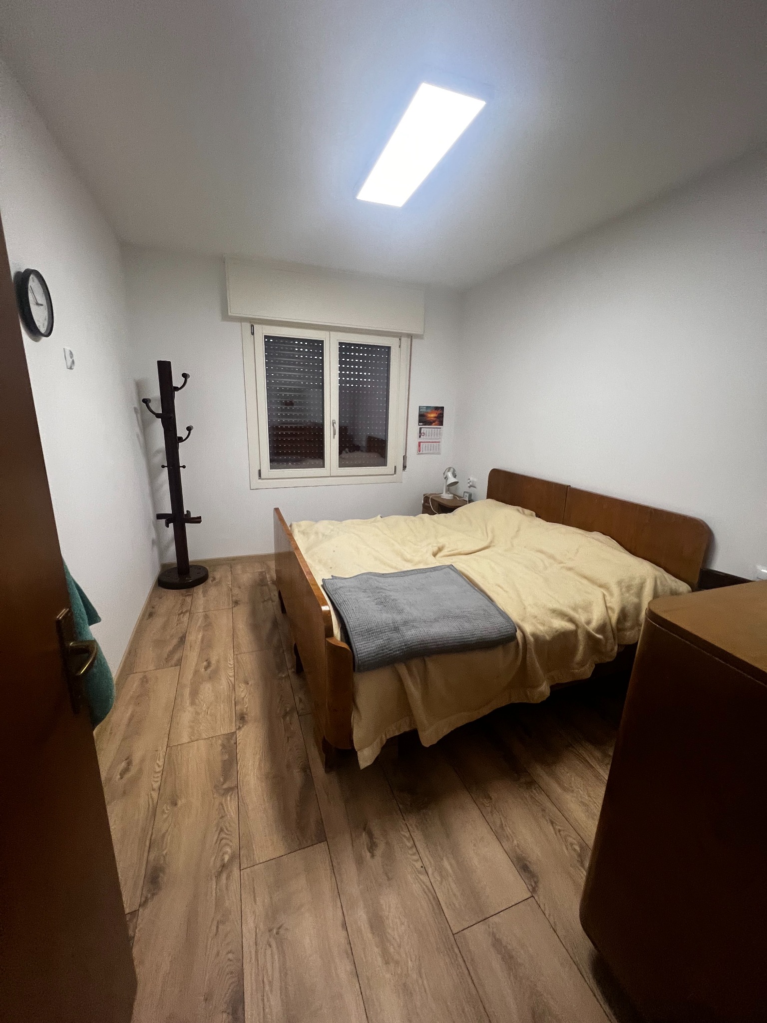 Jasna sypialnia po remoncie: drewniana podłoga, łóżko z beżową pościelą, w tle okno z żaluzjami i wieszak na ubrania przy ścianie. Sufit z oświetleniem.