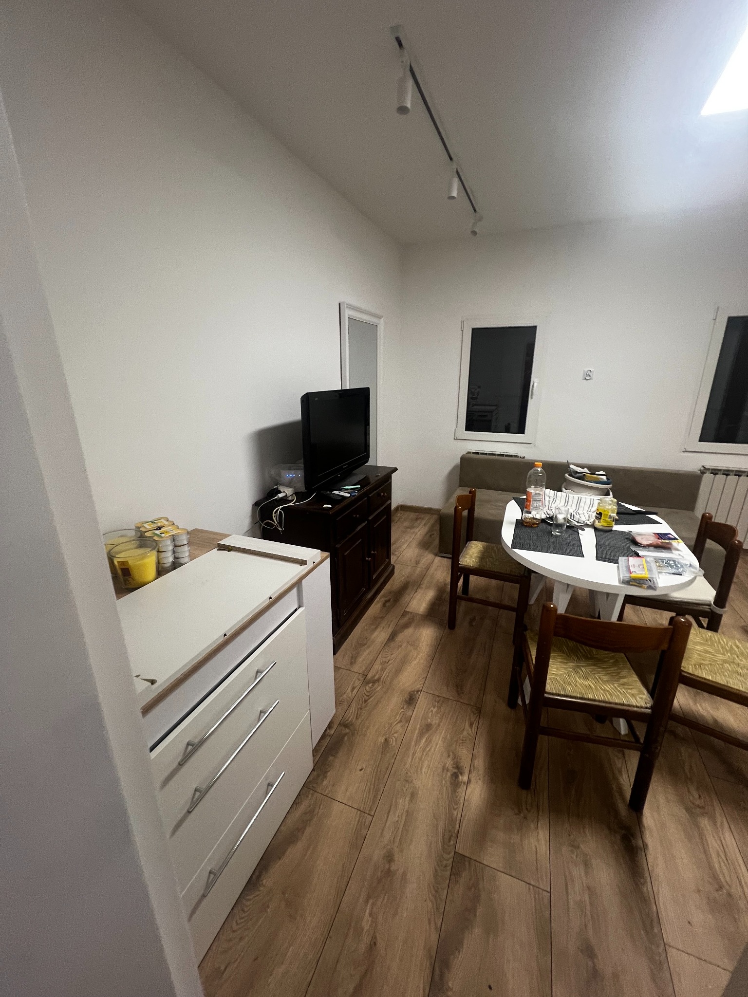 Jasne wnętrze po remoncie: drewniana podłoga, białe ściany, stół z krzesłami, komoda z telewizorem. Widoczne świeże wykończenia i oświetlenie sufitowe.