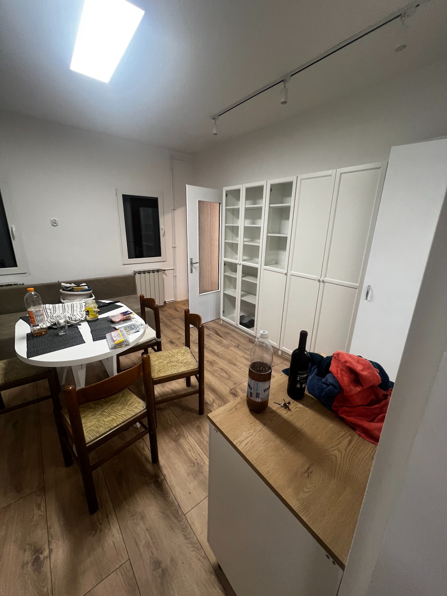 Jasne wnętrze po remoncie: drewniana podłoga, białe szafy wnękowe, stół z krzesłami. Widoczne ślady użytkowania, butelki i ubrania. Okno i oświetlenie sufitowe.
