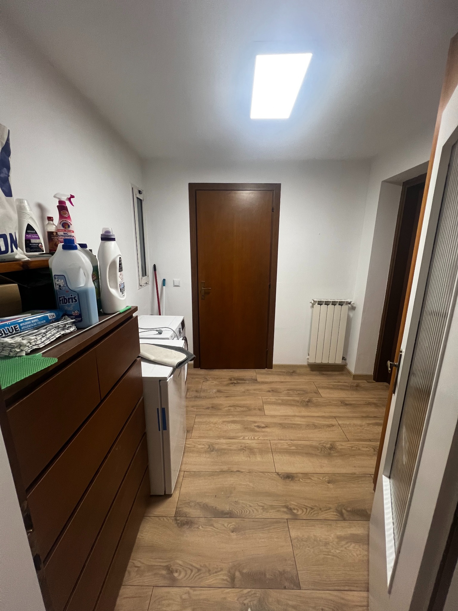 Wnętrze pomieszczenia gospodarczego z drewnianą podłogą, grzejnikiem i drzwiami. Widoczne środki czystości na komodzie i pralka. Oświetlenie sufitowe.
