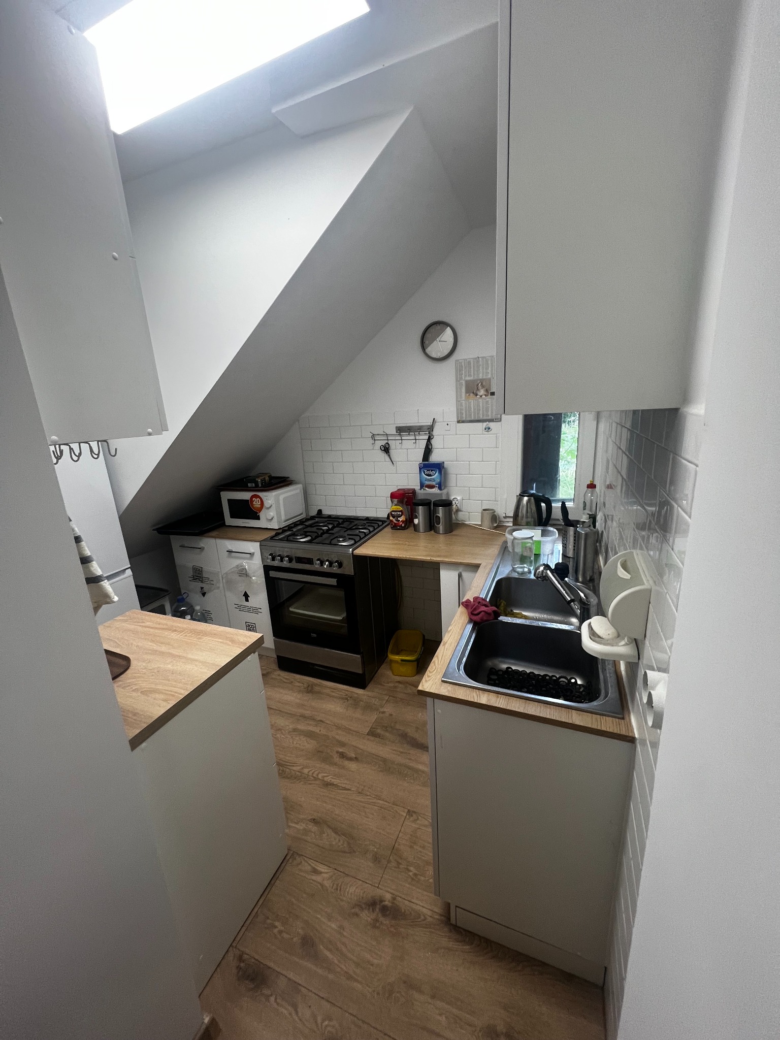 Kuchnia na poddaszu z białymi meblami, drewnianym blatem i płytkami na ścianie. Widoczny zlew, kuchenka i okno. Ustawiona pod skosem dachu, oświetlona światłem dziennym.