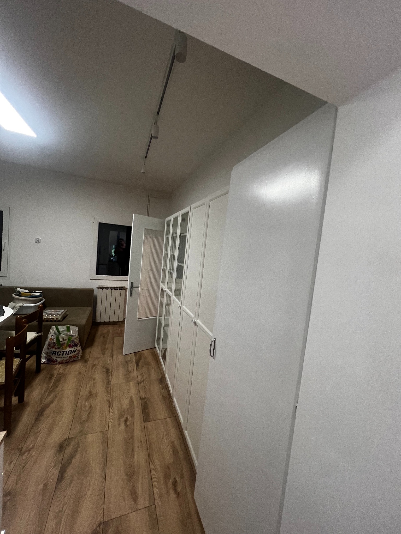 Białe szafy wnękowe z przeszklonymi drzwiami, widok na drewnianą podłogę i okno. Minimalistyczne wnętrze po remoncie, oświetlenie punktowe.