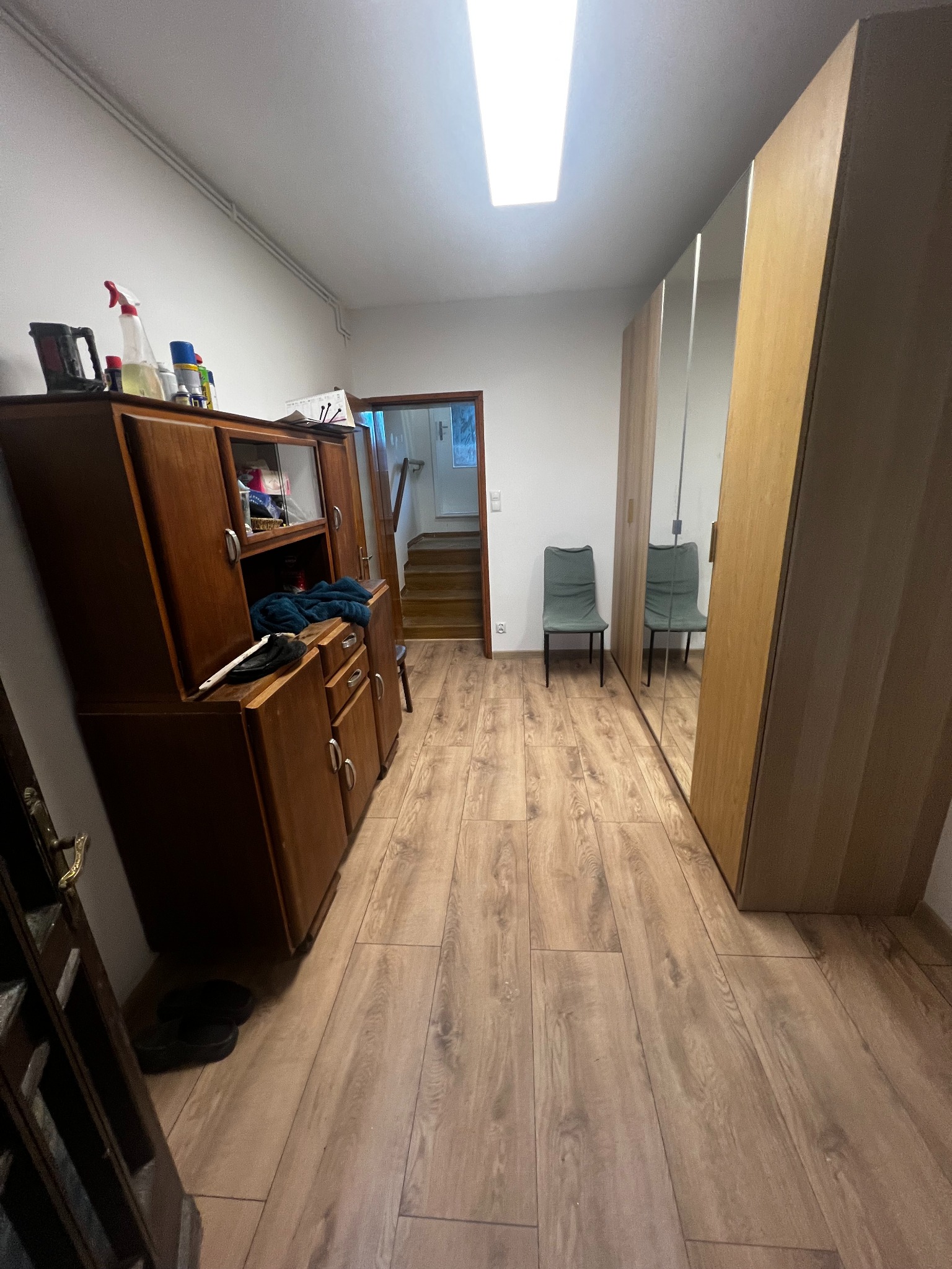Korytarz po remoncie: drewniana podłoga, szafa z lustrami, krzesło, stara komoda. Widok na schody.  Światło sufitowe. Buty przy drzwiach.