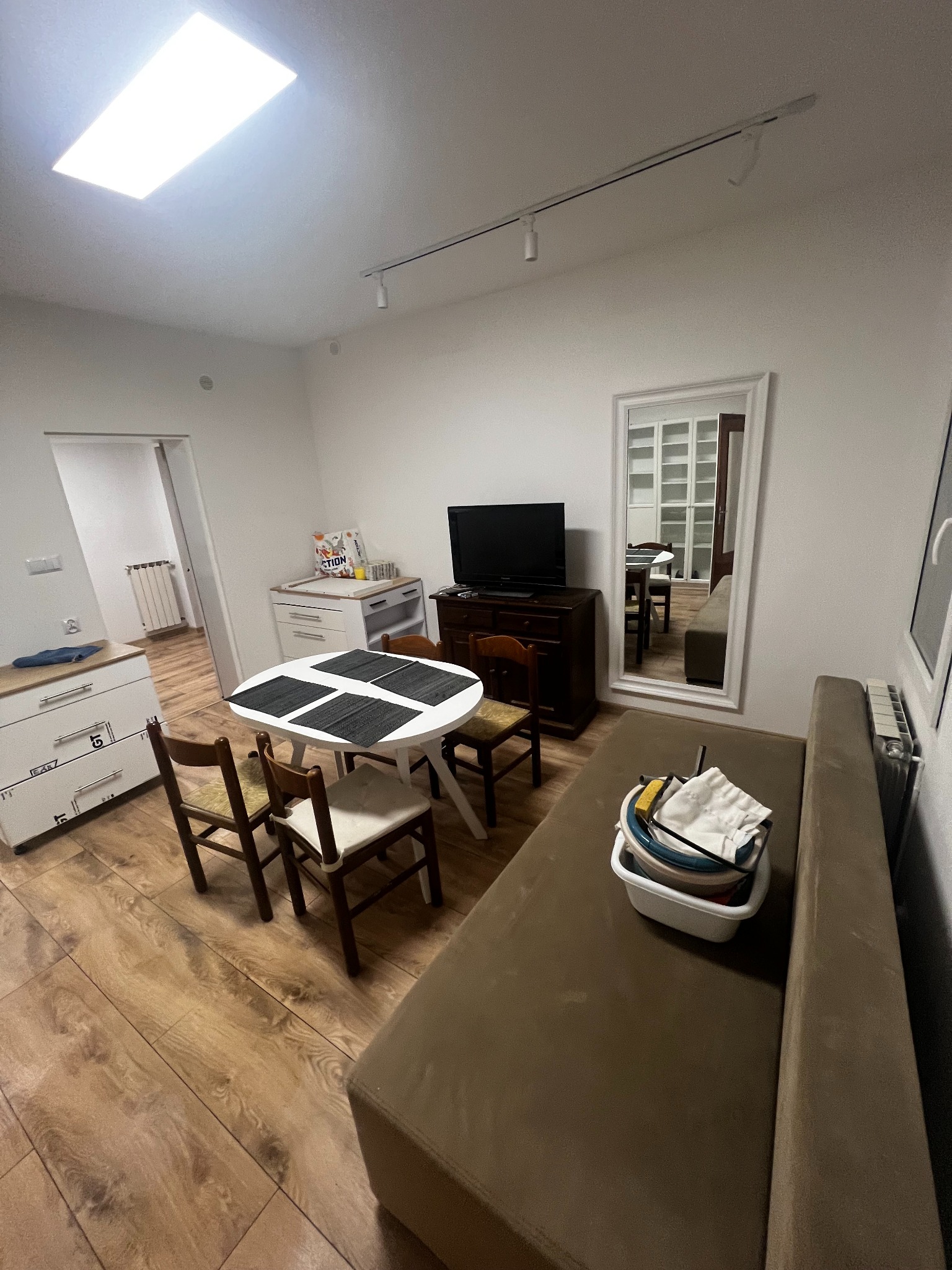 Jasne wnętrze po remoncie: drewniana podłoga, białe meble, stół z krzesłami i sofa. W tle lustro odbija dalszą część pomieszczenia, a na suficie widoczne oświetlenie.