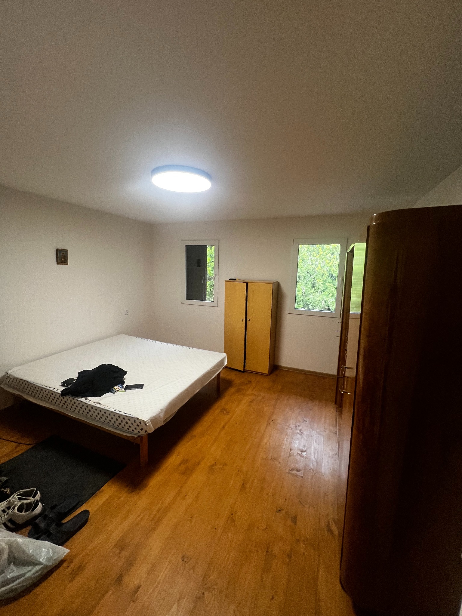 Pokój po remoncie: drewniana podłoga, białe ściany, okna z widokiem na zieleń. Łóżko z materacem i garderoba. Minimalistyczny design, jasne oświetlenie.