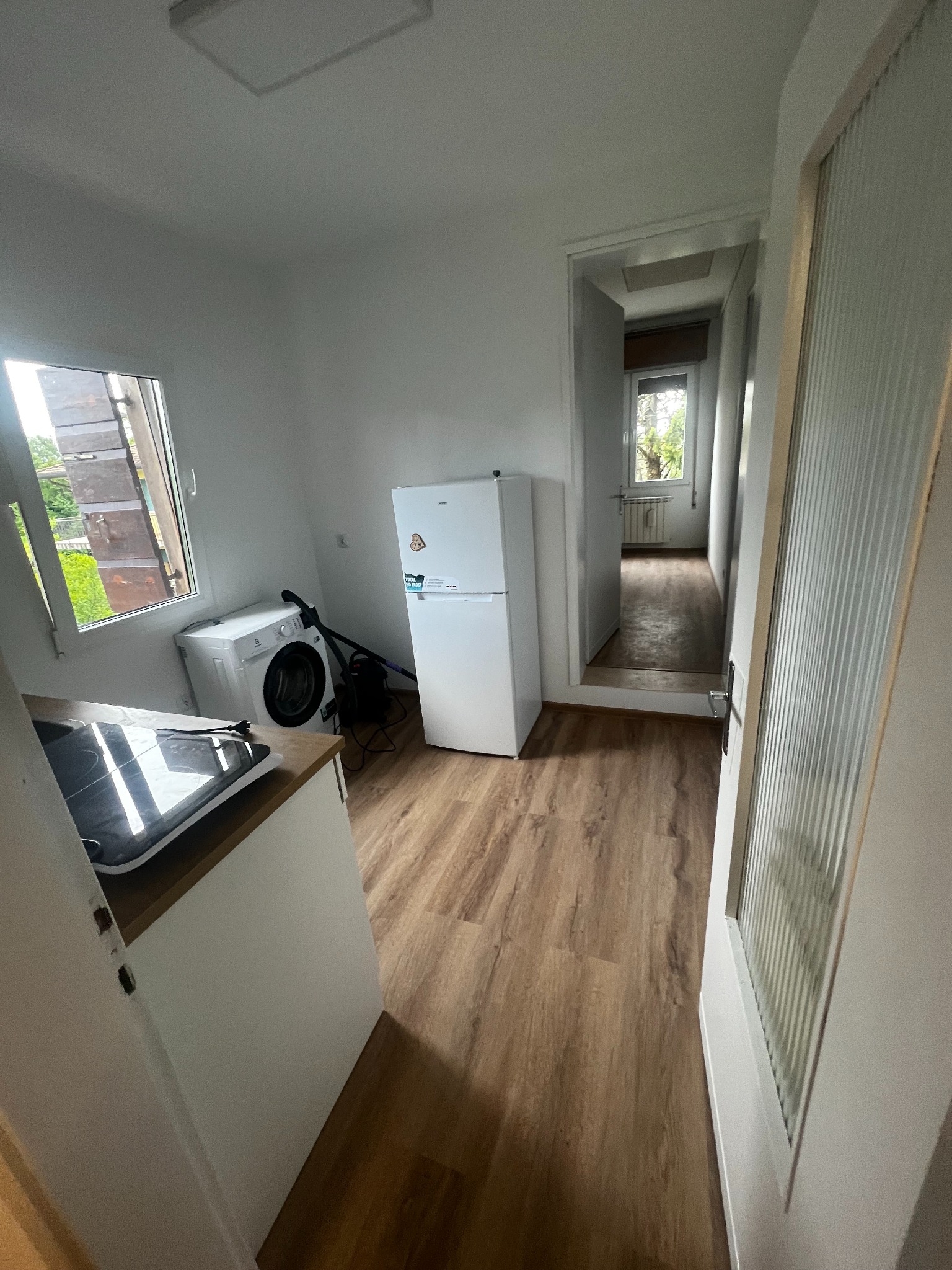 Wykończona kuchnia z drewnianą podłogą: biała zabudowa kuchenna, okno z widokiem na zieleń, pralka, lodówka. Widok na dalsze pomieszczenie przez otwarte drzwi.