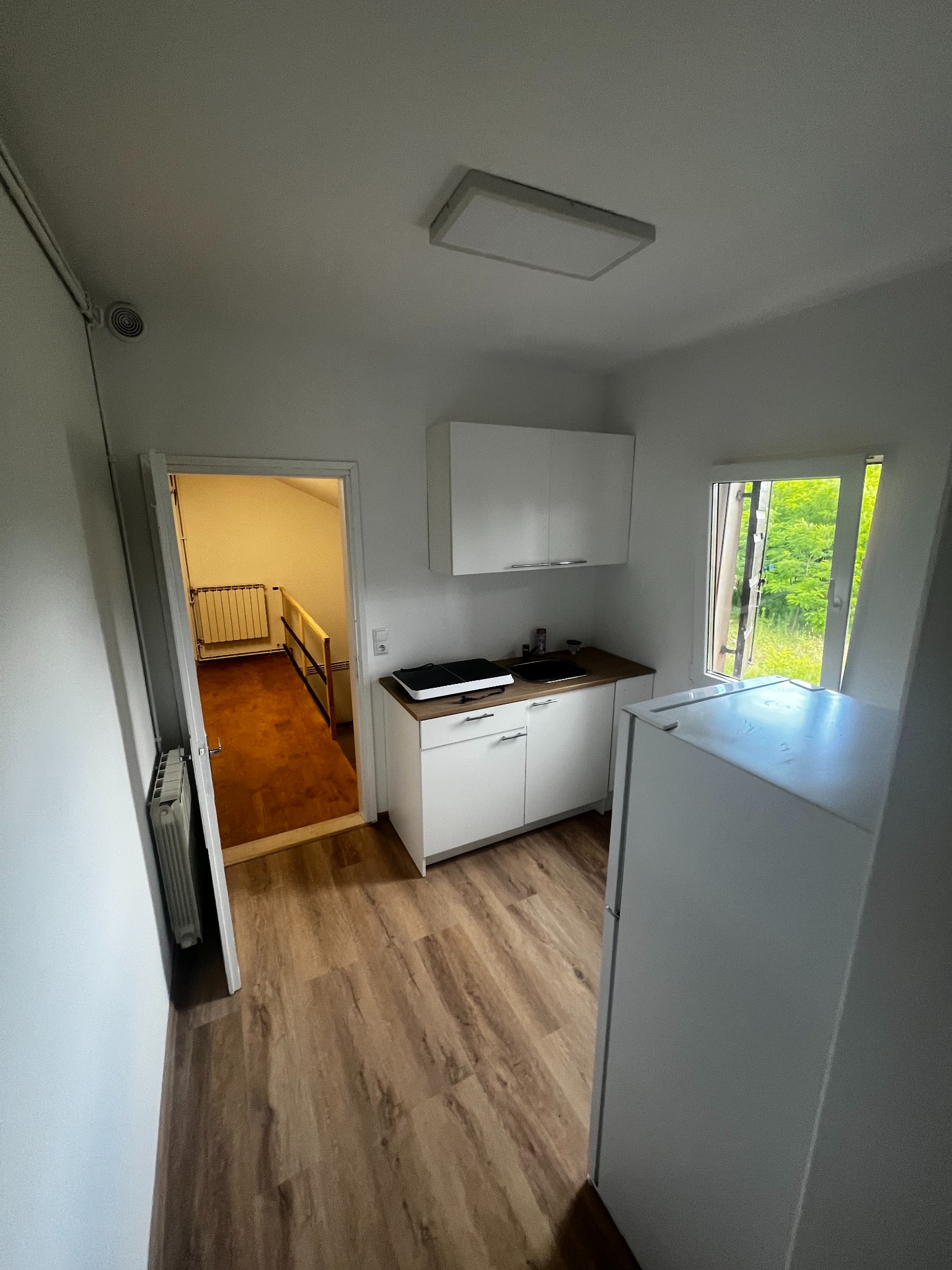 Biała, minimalistyczna kuchnia z drewnianą podłogą i widokiem na schody. W tle zieleń za oknem. Proste szafki, lodówka i oświetlenie sufitowe.