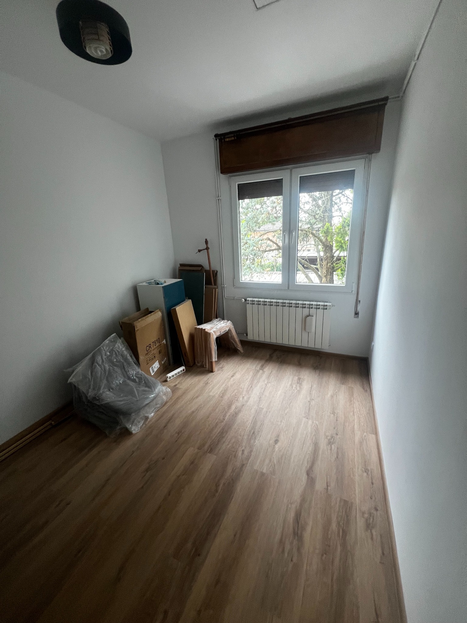 Pusta, jasna przestrzeń z panelami podłogowymi. Okno z grzejnikiem, kartony i meble pod ścianą. Przygotowanie do remontu, widok na zewnątrz.