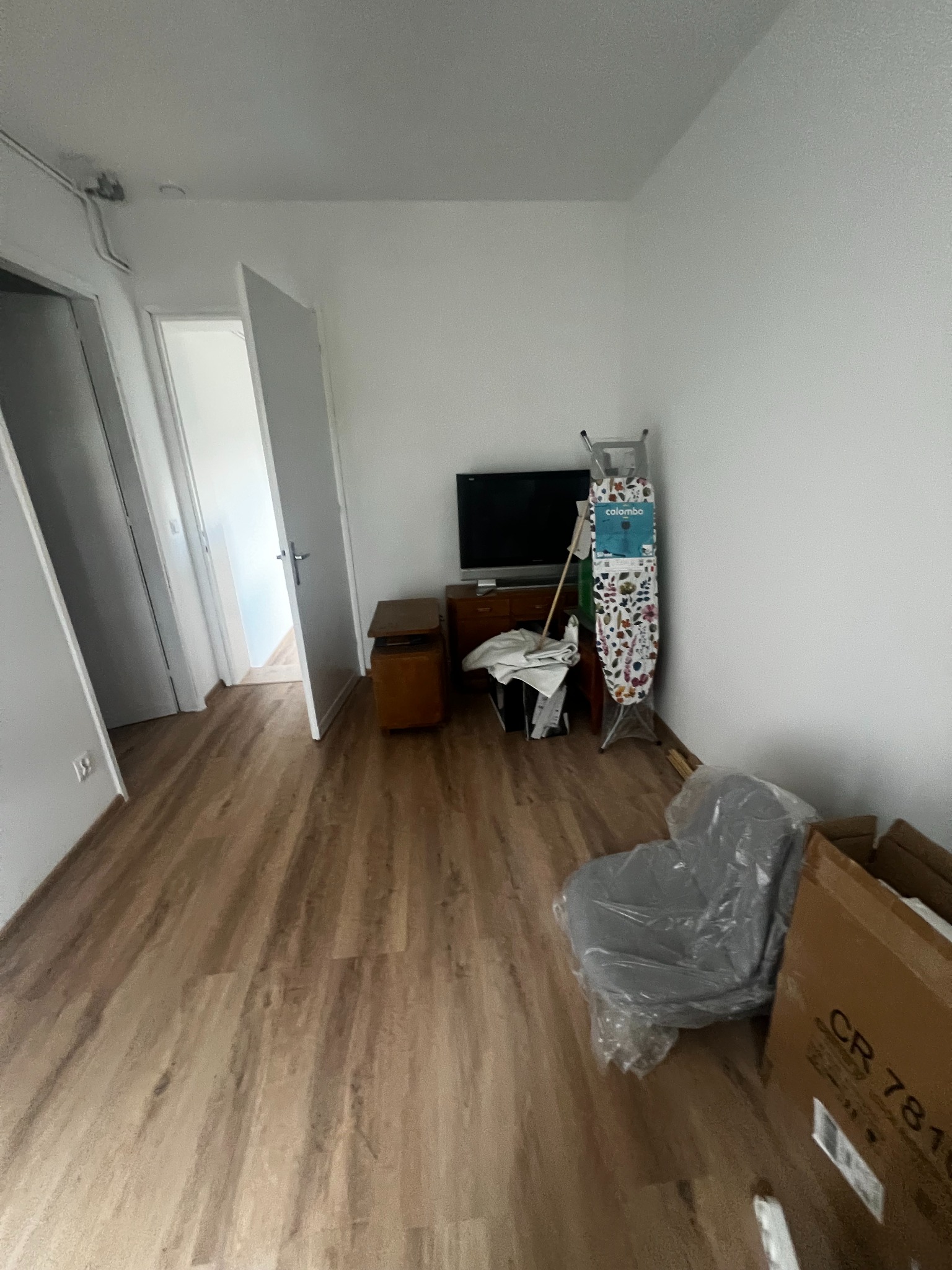 Wnętrze pokoju z panelami podłogowymi i otwartymi drzwiami. Widoczny telewizor, deska do prasowania i krzesło w folii. Przygotowania do remontu.