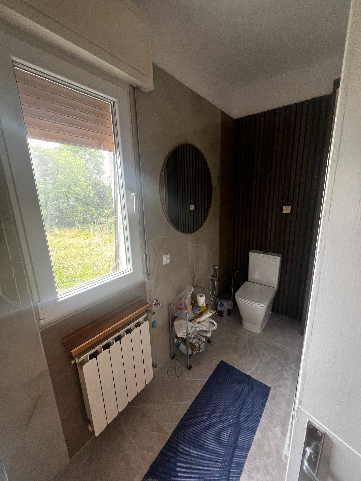 Wykończona łazienka z oknem na zewnątrz, szare płytki, drewniane panele na ścianie, okrągłe lustro i biała toaleta. Widoczny także grzejnik pod oknem.