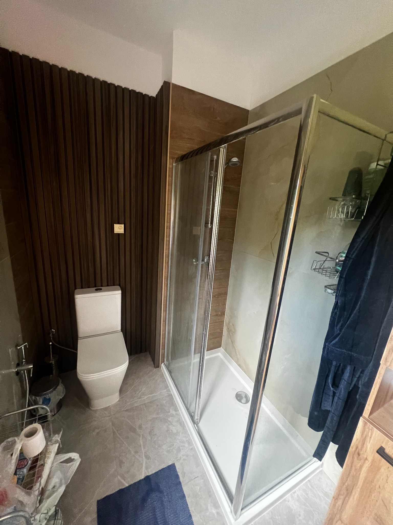 Nowoczesna łazienka po remoncie: kabina prysznicowa ze srebrnymi ramami, toaleta i dekoracyjne panele ścienne w odcieniach brązu. Szare płytki podłogowe.