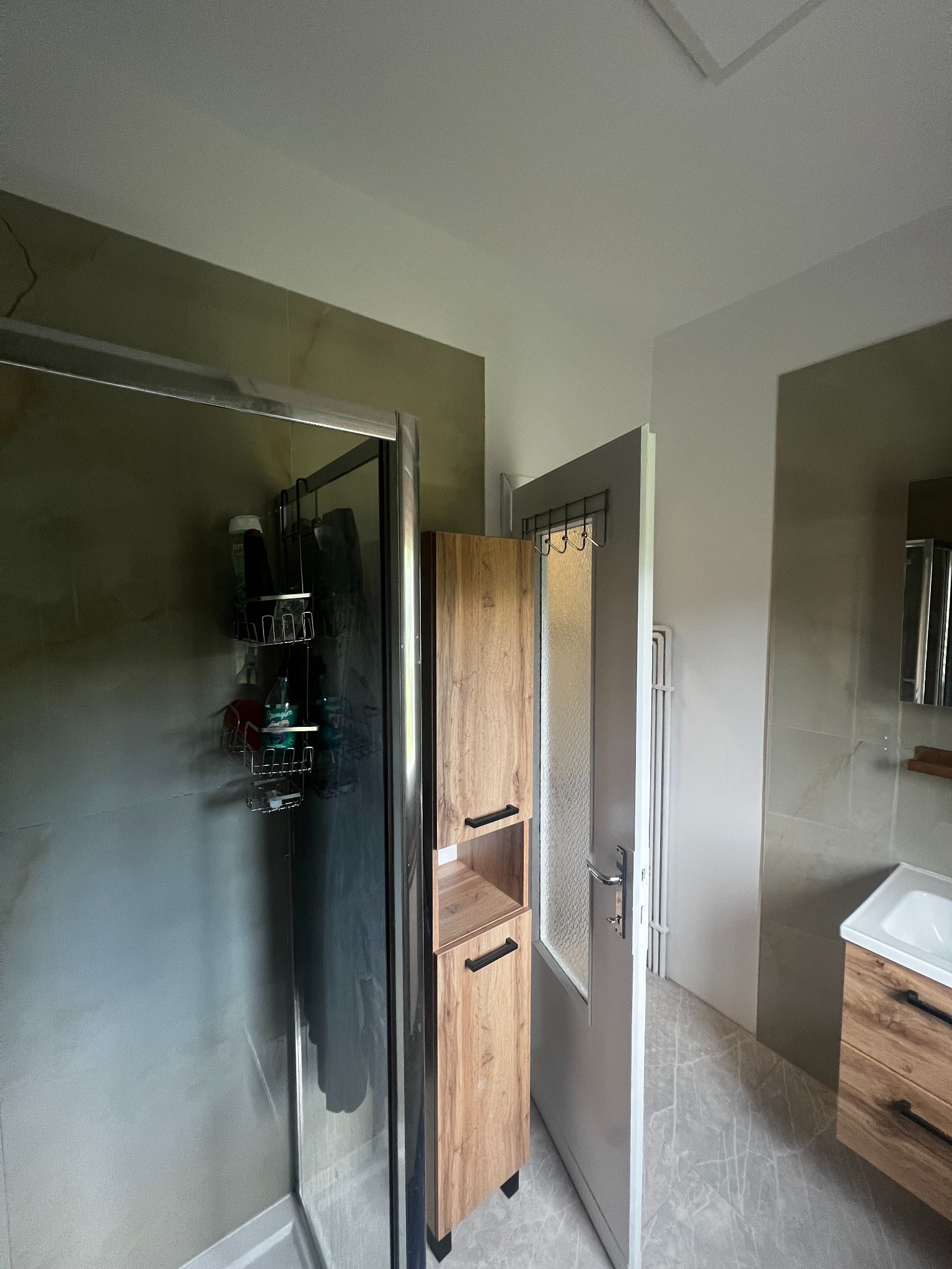 Wykończona łazienka w odcieniach szarości i drewna: kabina prysznicowa, szafka z drewnianym fornirem, umywalka i drzwi z wieszakami. Widok na wnętrze po remoncie.
