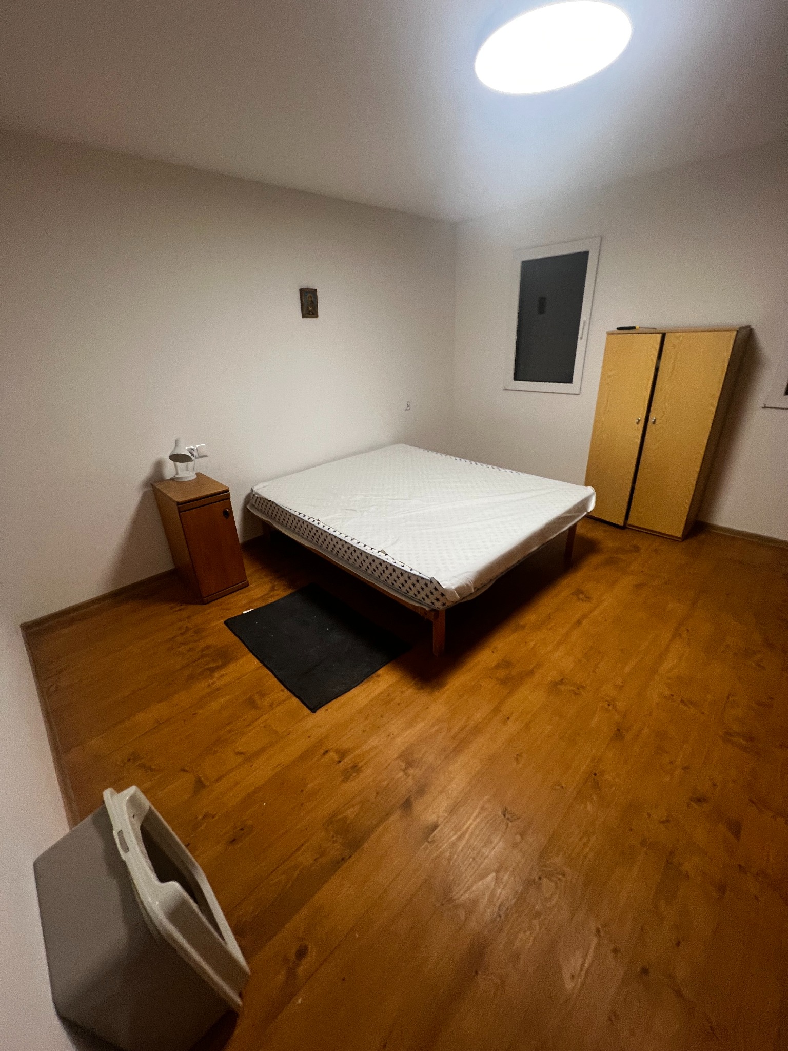 Pokój z drewnianą podłogą, łóżkiem z białym materacem, szafką nocną i szafą. Widoczne okno i lampa sufitowa. Dom po remoncie.