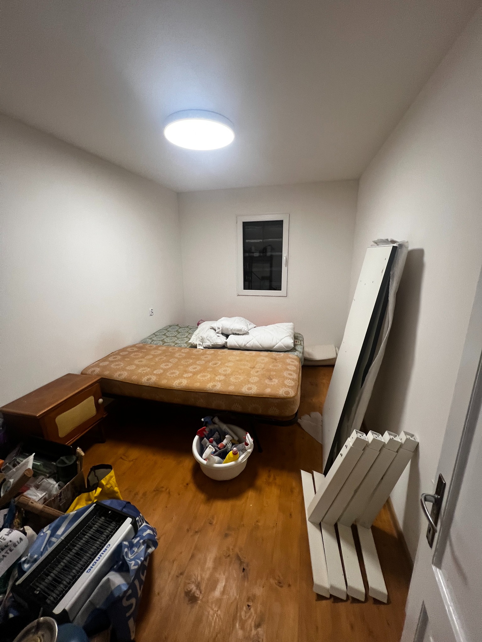 Pokój w trakcie remontu: łóżko, okno, białe ściany, podłoga z desek, elementy wyposażenia i narzędzia. Przygotowanie do prac wykończeniowych.