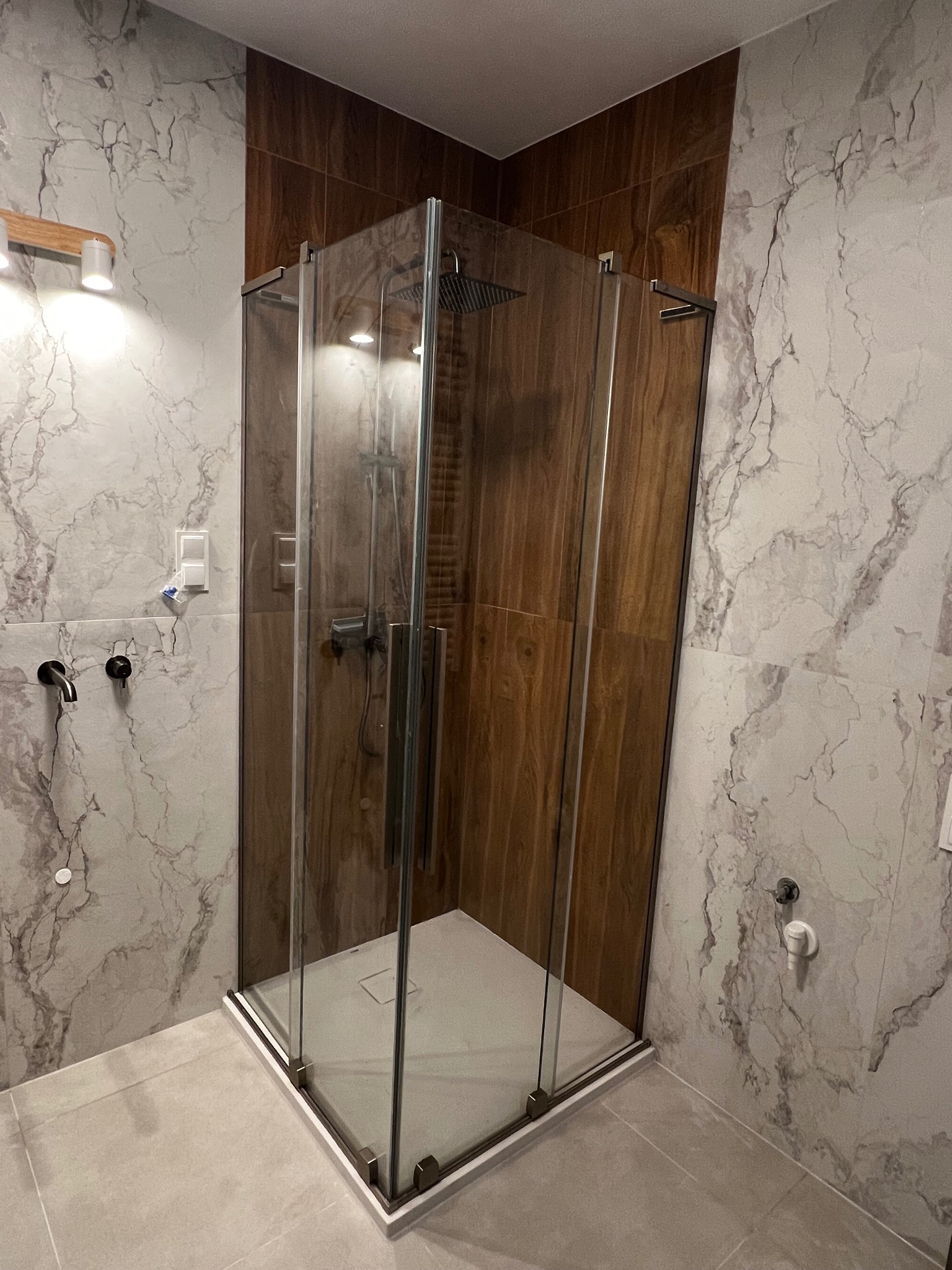 Nowoczesna kabina prysznicowa z deszczownicą, obudowana płytkami imitującymi marmur i drewno, w jasnej łazience. Widoczne detale wykończenia i armatura.