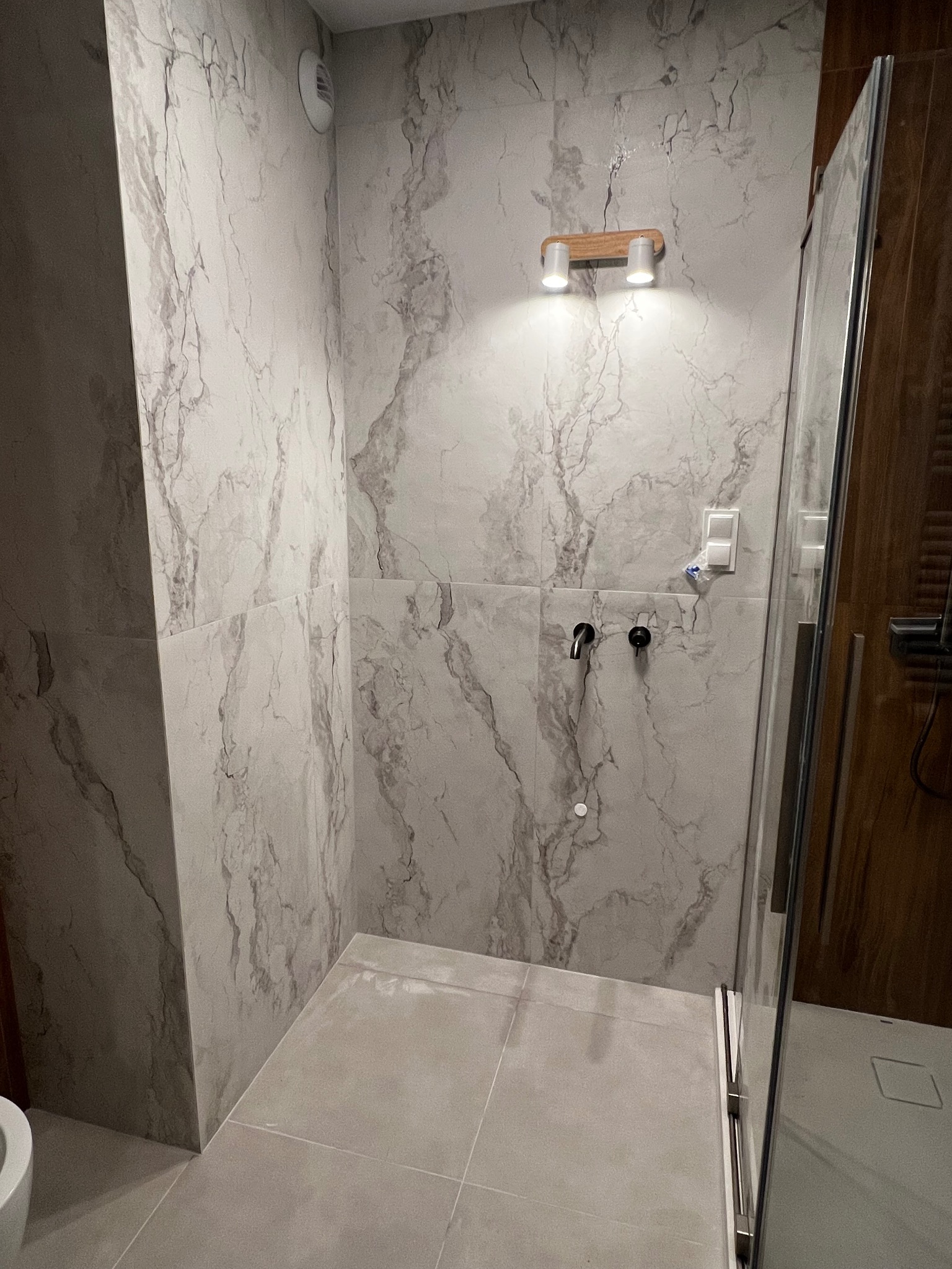 Wykończona kabina prysznicowa z marmurowymi płytkami, oświetleniem punktowym i szklanymi drzwiami. Widoczne baterie i odpływ liniowy. Minimalistyczny design.