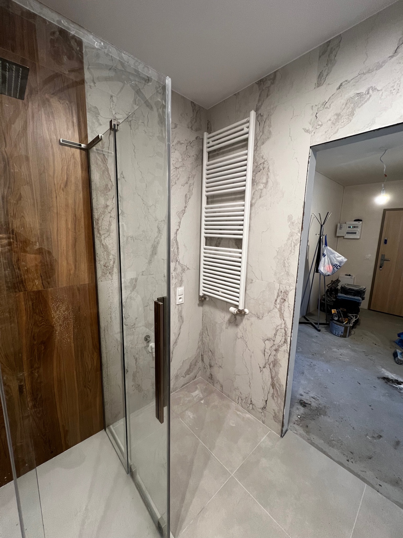 Wykończona łazienka z kabiną prysznicową typu walk-in, grzejnikiem drabinkowym i płytkami imitującymi marmur. Widok na pomieszczenie w trakcie prac wykończeniowych.