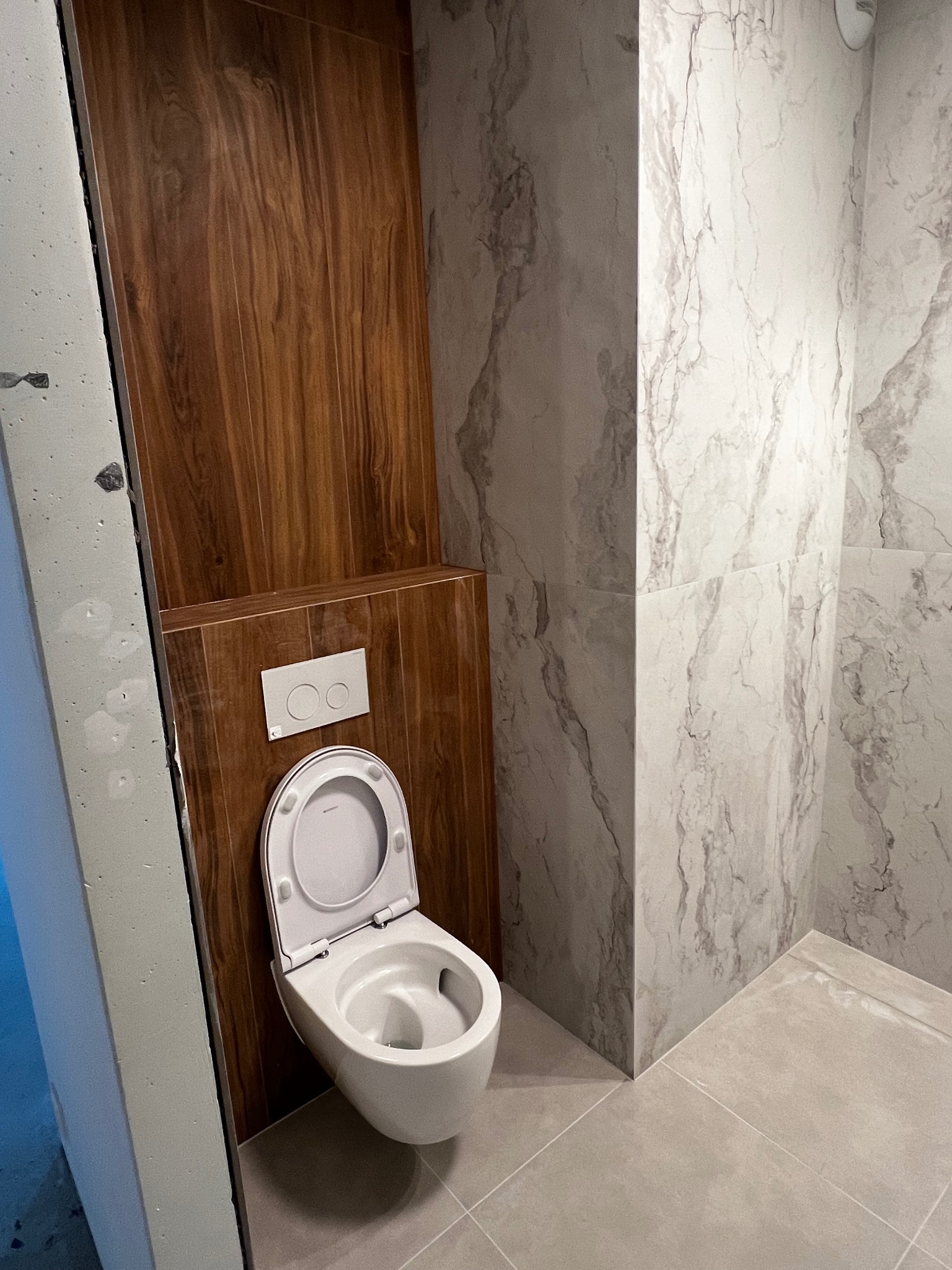 Nowoczesna toaleta podwieszana w łazience wykończonej płytkami imitującymi drewno i marmur. Widoczny fragment ściany z przyciskiem spłukującym.