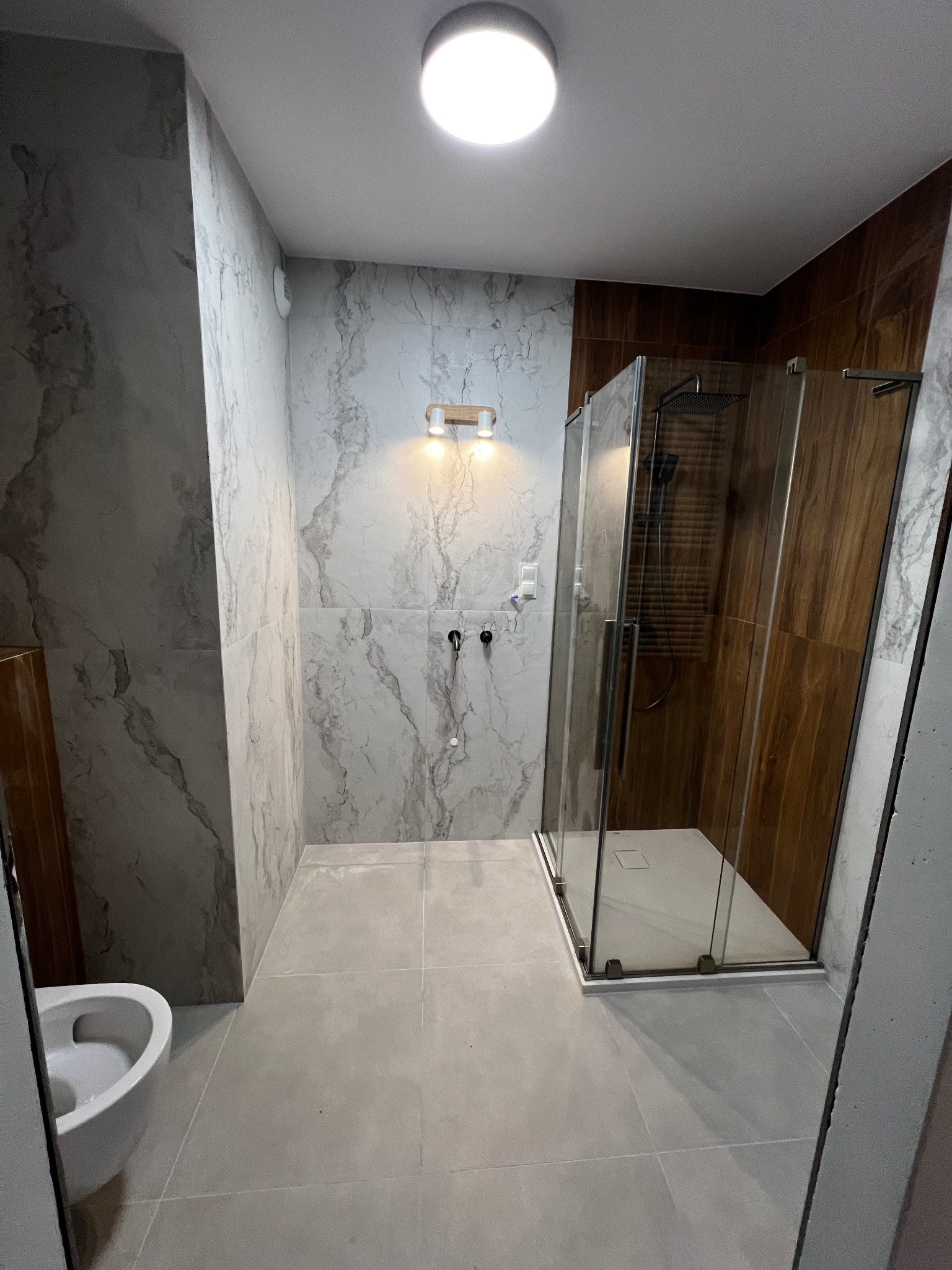 Nowoczesna łazienka po remoncie: kabina prysznicowa ze szklanymi drzwiami, marmurowe płytki na ścianach, szara podłoga i wisząca toaleta. Minimalistyczny design.