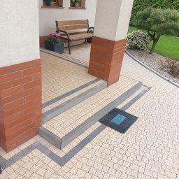 KJB cleaning - Ułożona kostka brukowa w odcieniach beżu i szarości, tworząca wzór na podjeździe. Widoczne schody wejściowe i fragment kolumny domu. Mała wycieraczka przy schodach.