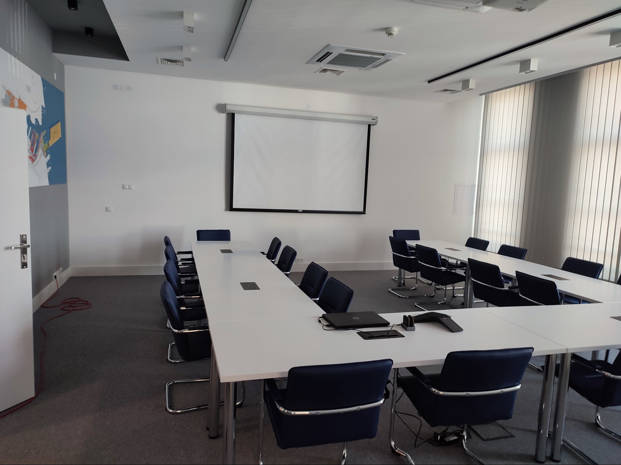 Jasne, przestronne biuro z długim stołem konferencyjnym w kształcie litery U, otoczonym przez ciemnoniebieskie krzesła biurowe na metalowych nogach, z widocznym rzutnikiem i ekranem na ścianie...