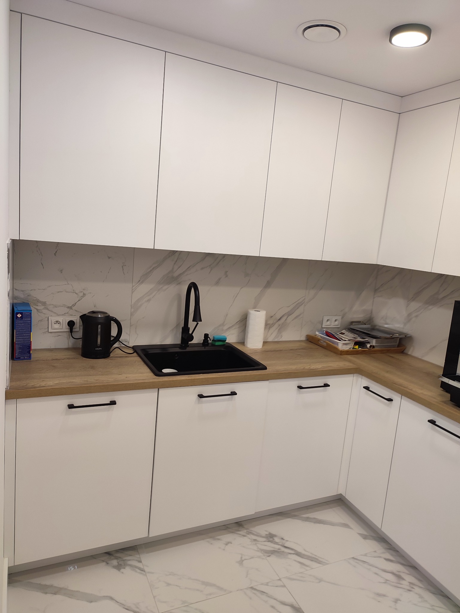 Nowoczesna kuchnia z białymi, minimalistycznymi szafkami, drewnianym blatem i czarną armaturą, uzupełniona marmurkową płytką na ścianie i podłodze.