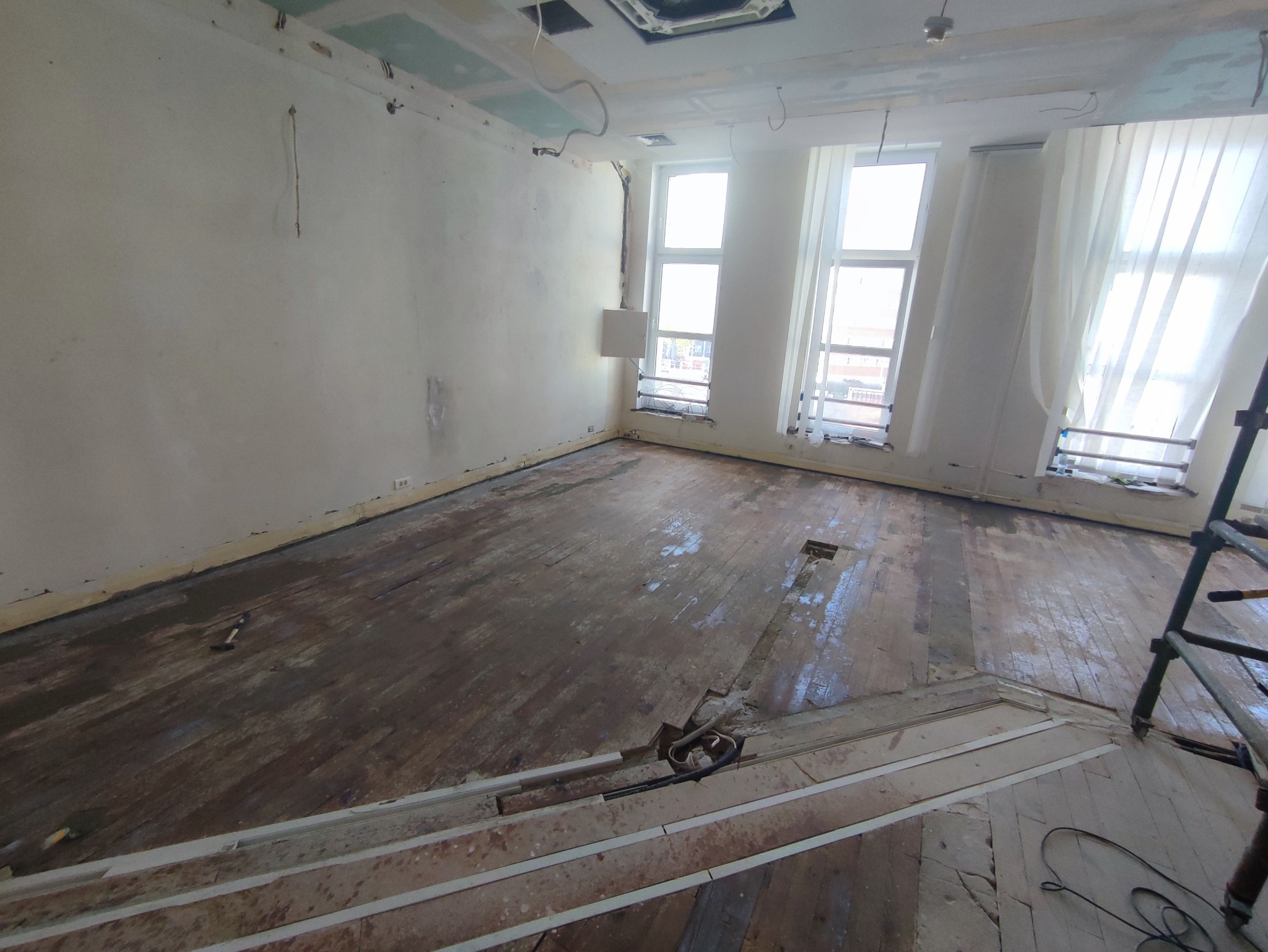 Wnętrze pokoju w trakcie remontu: surowe ściany z widocznymi śladami po instalacjach, odsłonięta drewniana podłoga z fragmentami usuniętych desek, okna z białymi zasłonami przepuszczającymi jasne...