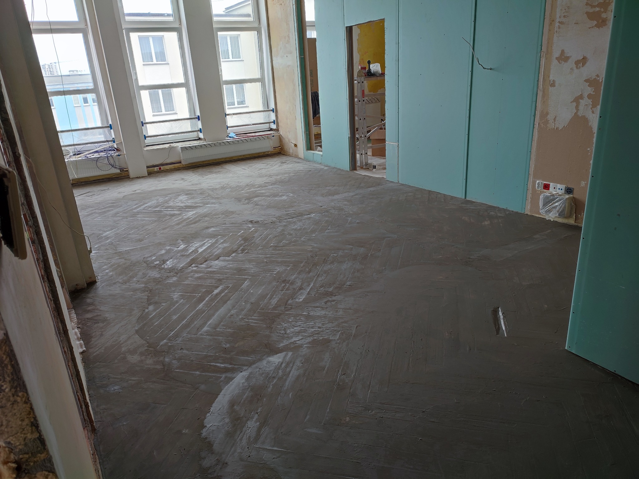 Przygotowanie podłoża pod parkiet w jodełkę w trakcie remontu, widoczne ślady szpachli na podłodze, ściany częściowo pokryte płytami gipsowo-kartonowymi w kolorze turkusowym, okna z widokiem...