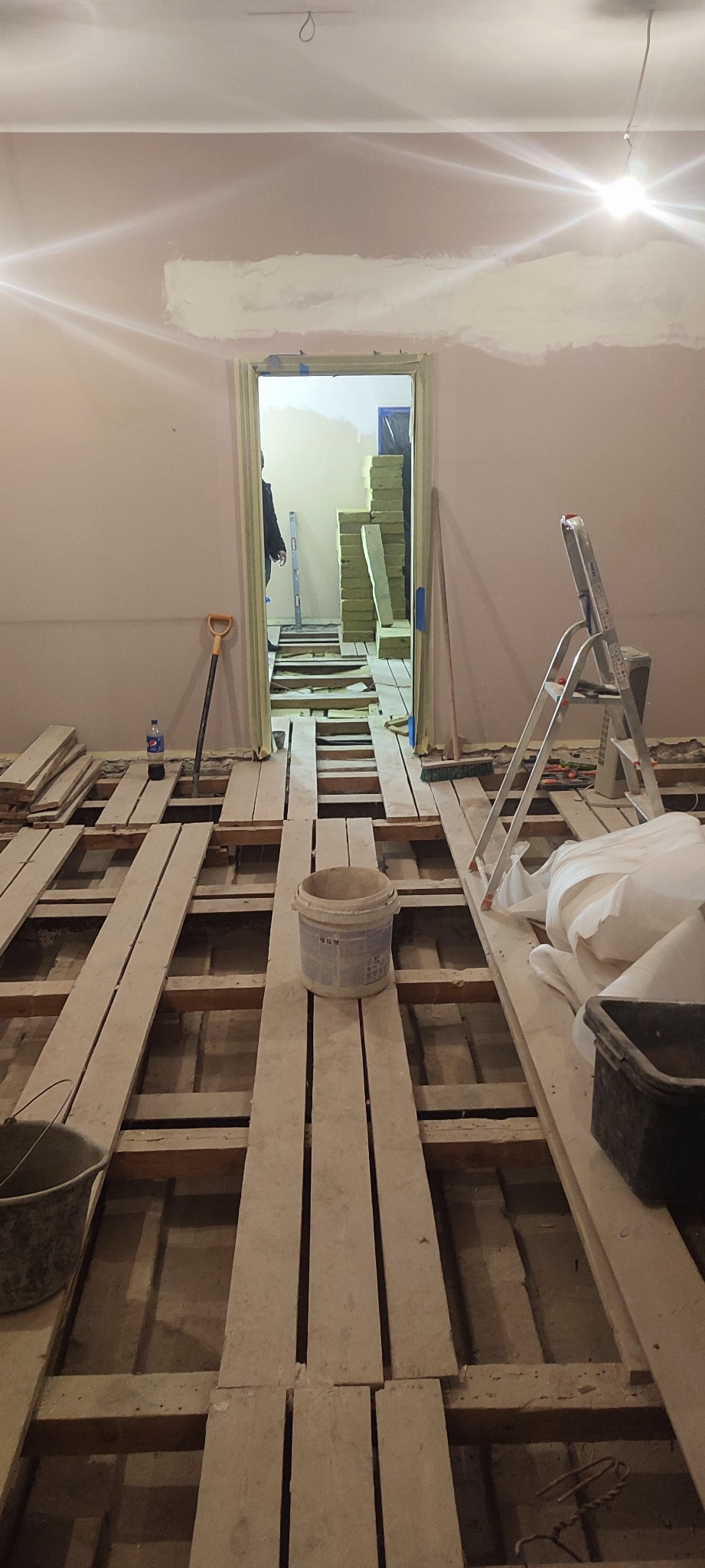 Remontowany pokój z odsłoniętą konstrukcją podłogi, widoczne belki, deski, wiadro z farbą, drabina, oraz sterta materiałów izolacyjnych w przejściu do sąsiedniego pomieszczenia, ściany częściowo...