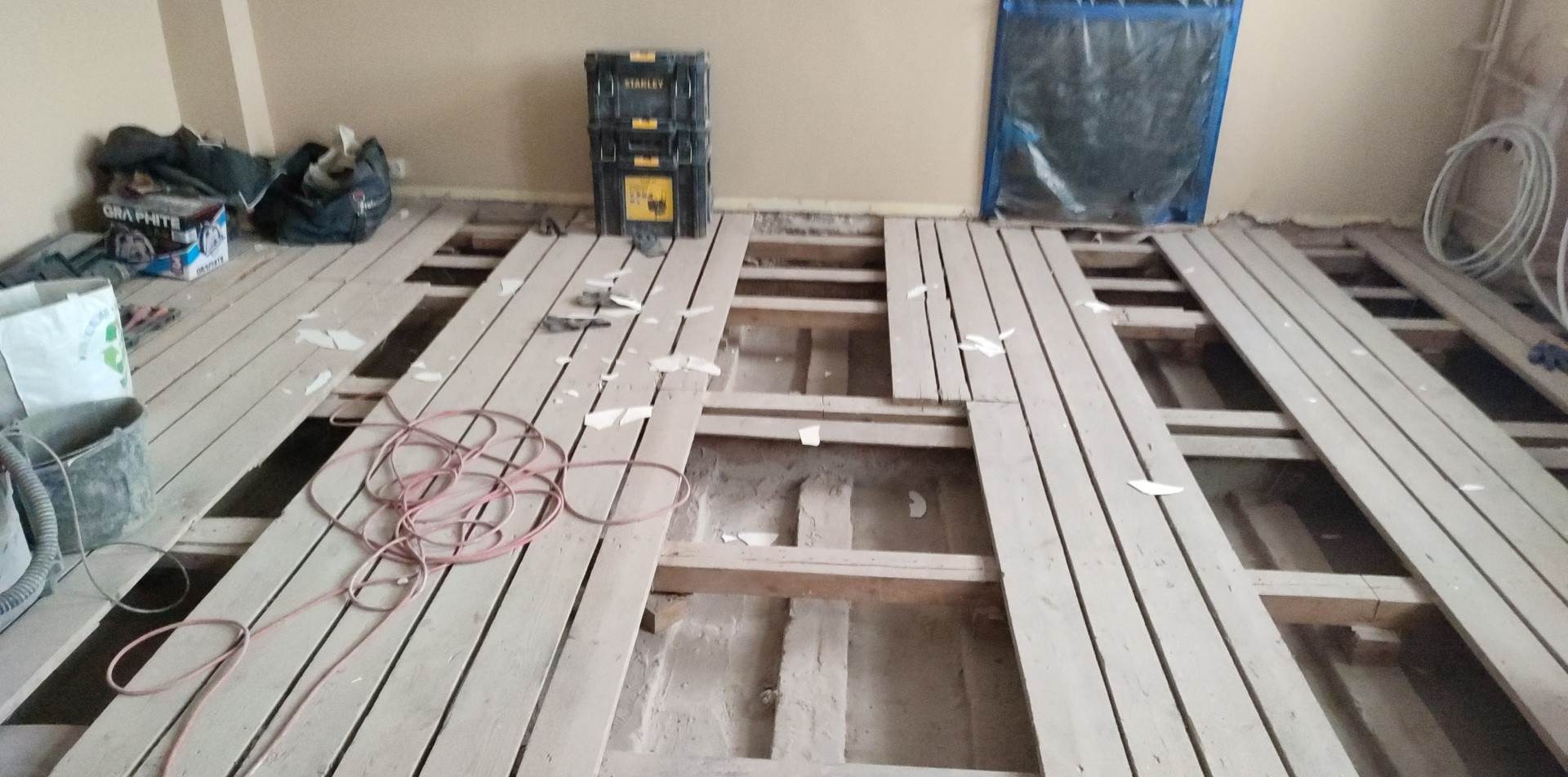 Demontaż starej podłogi drewnianej w trakcie remontu, widoczne deski podłogowe z przerwami, narzędzia i materiały budowlane w tle, okno zabezpieczone folią.