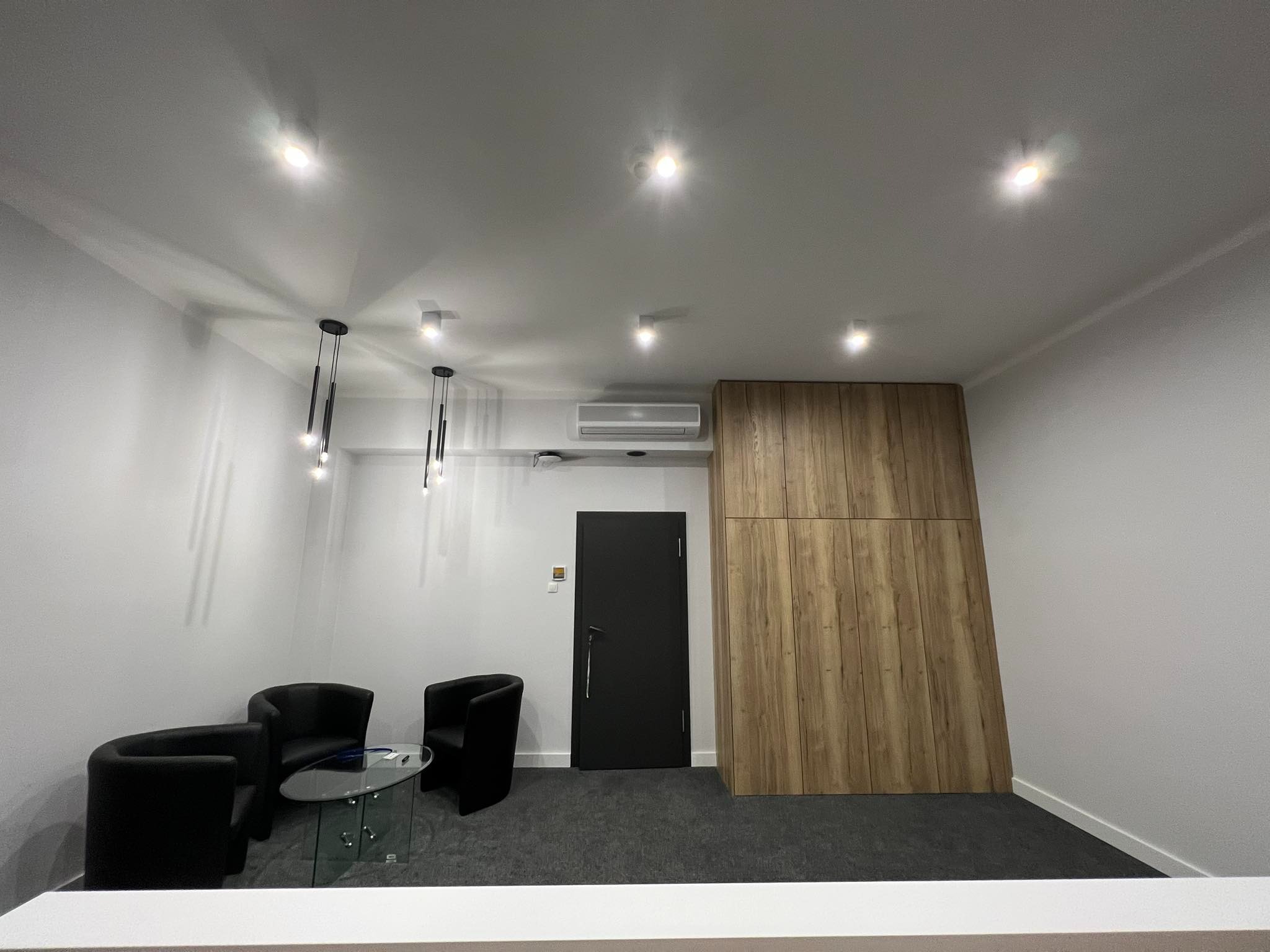 Nowoczesne wnętrze biurowe z czarnymi fotelami klubowymi wokół szklanego stolika, minimalistycznymi lampami wiszącymi i zabudową ściany z paneli drewnopodobnych, oświetlone punktowymi lampami...