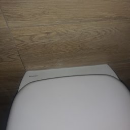 Robert Śniadach - Zamontowana biała toaleta marki Geberit na tle drewnopodobnych płytek ściennych, widoczne niewielkie ślady silikonu.