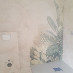 Robert Śniadach - Wykończona łazienka z beżowymi płytkami imitującymi kamień, fragment ściany z motywem roślinnym, zamontowany stelaż podtynkowy WC i brodzik z odpływem liniowym.