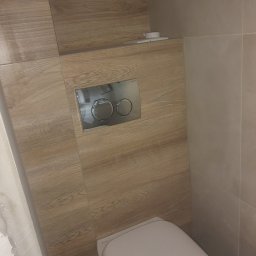 Robert Śniadach - Zamontowana toaleta podwieszana z chromowanym przyciskiem spłukującym na ścianie wykończonej płytkami imitującymi drewno, widoczne niedokończone prace na ścianie po lewej stronie.