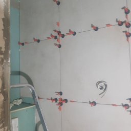Robert Śniadach - Ściana w trakcie układania płytek, widoczne systemy poziomowania płytek (klipsy i kliny), fragment drabiny i surowa ściana obok, przygotowywana instalacja elektryczna.