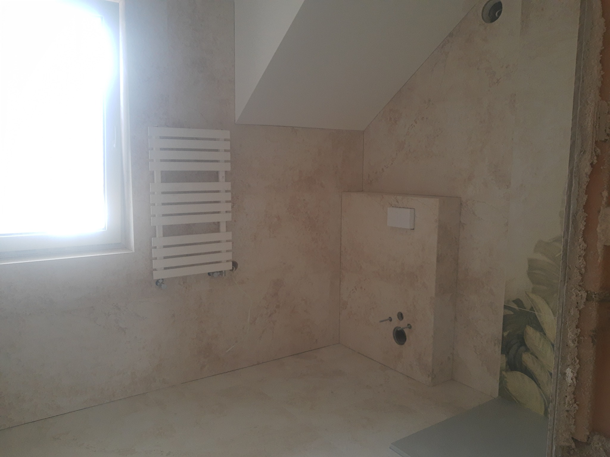 Jasna łazienka w trakcie wykańczania: beżowe płytki imitujące kamień, grzejnik drabinkowy przy oknie, zabudowa podtynkowego WC, widoczne przyłącza hydrauliczne i fragment dekoracyjnej płytki...