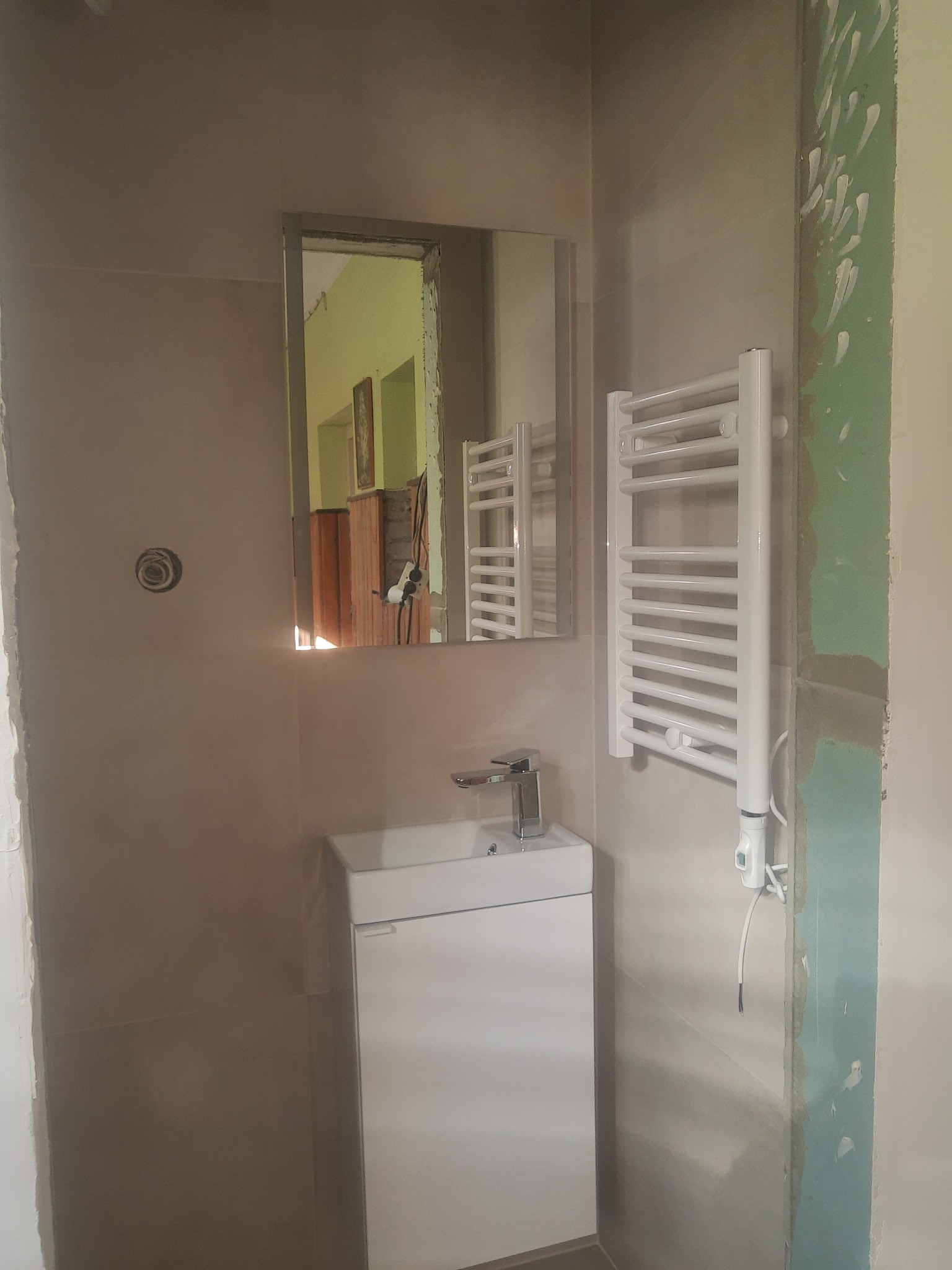 Mała łazienka w trakcie remontu: zamontowana biała umywalka z szafką, grzejnik drabinkowy i lustro, widoczne ślady po gładzi na ścianie.