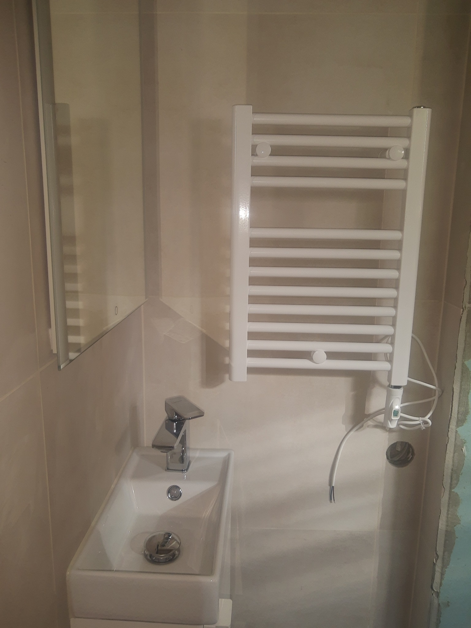 Mała łazienka w trakcie remontu: biała umywalka z chromowaną baterią, lustro z widocznym odbiciem, biały grzejnik drabinkowy elektryczny z widocznym kablem, ściany wyłożone jasnymi płytkami...