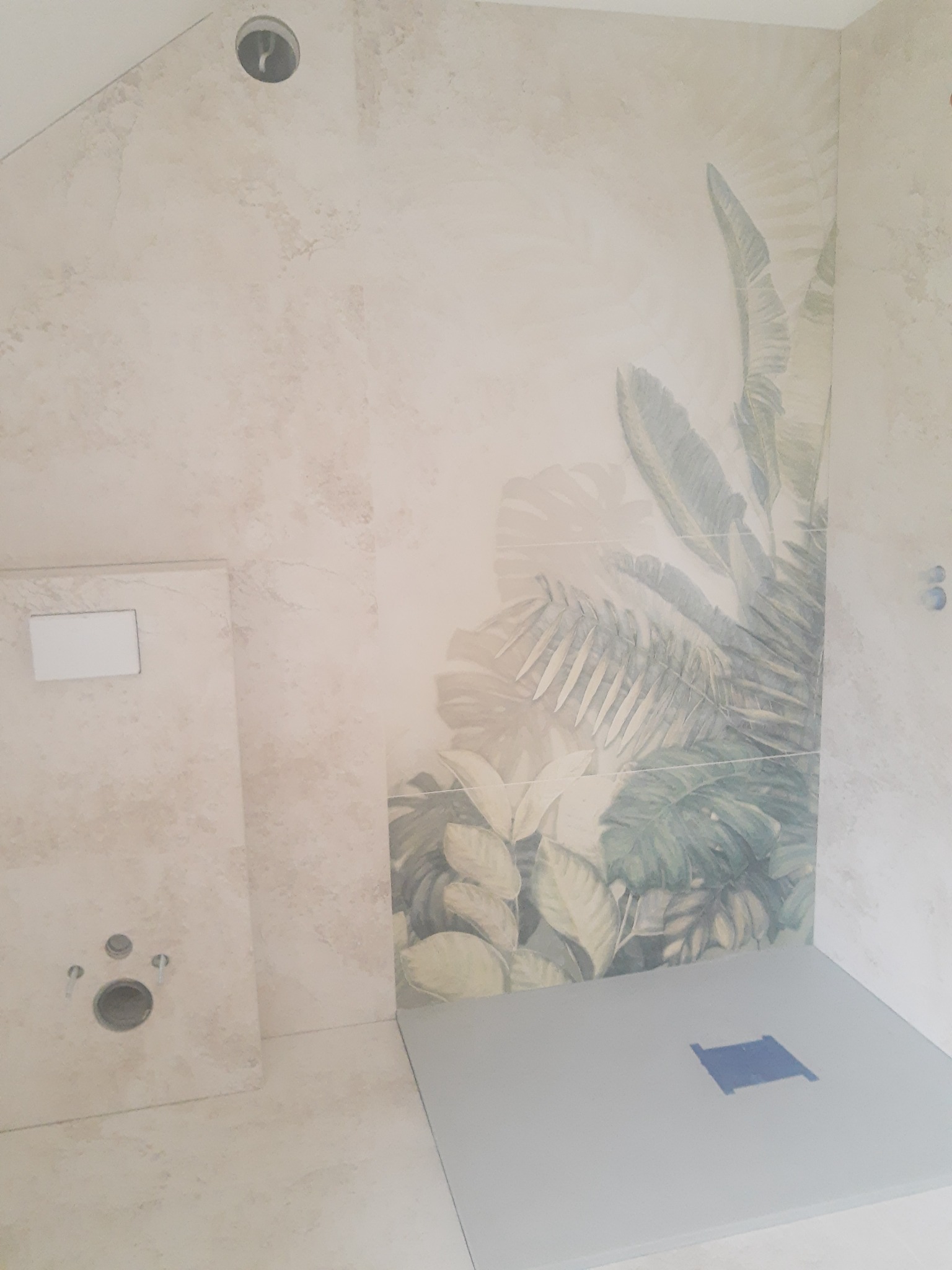 Wykończona łazienka z beżowymi płytkami imitującymi kamień, fragment ściany z motywem roślinnym, zamontowany stelaż podtynkowy WC i brodzik z odpływem liniowym.