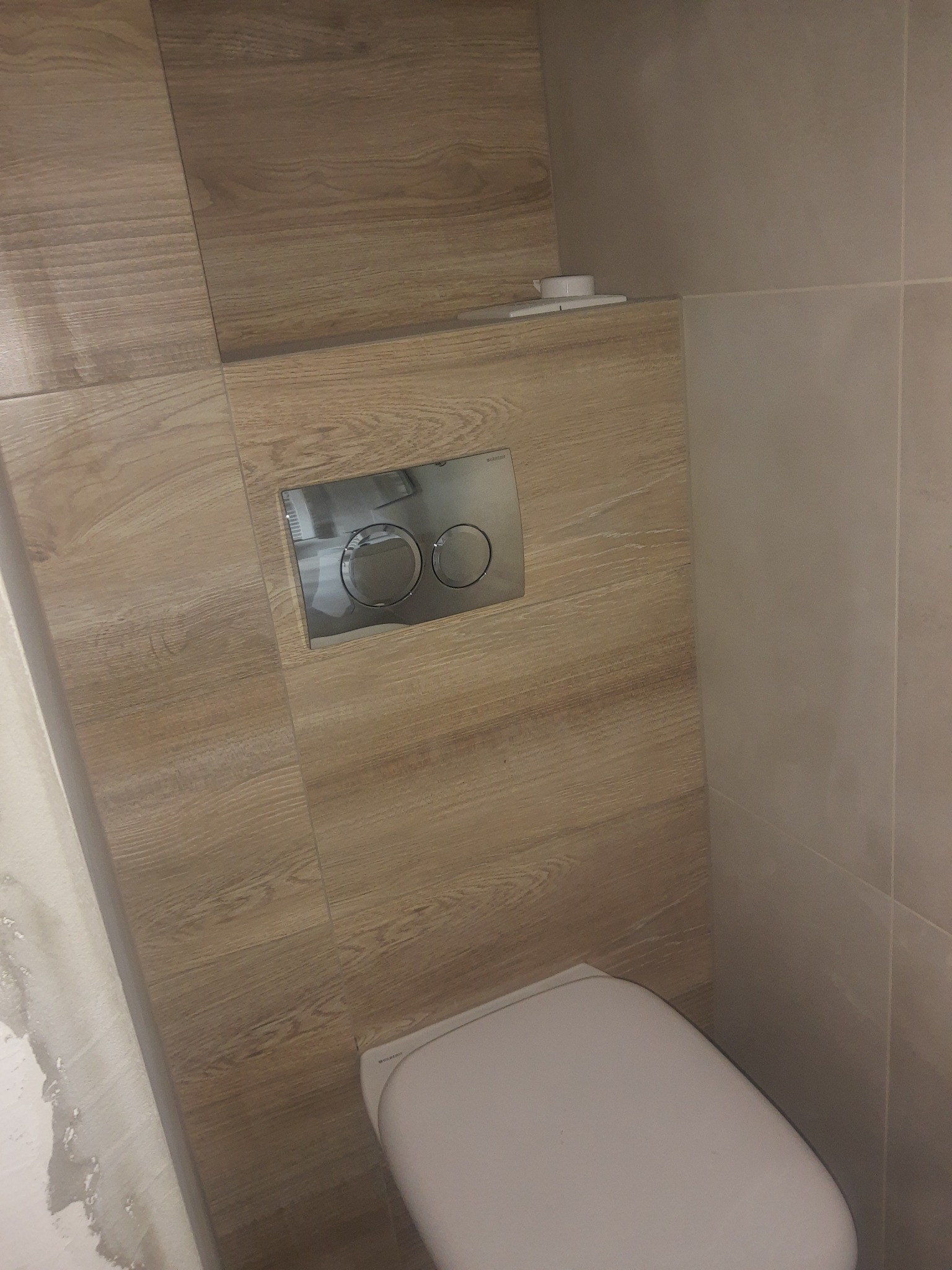 Zamontowana toaleta podwieszana z chromowanym przyciskiem spłukującym na ścianie wykończonej płytkami imitującymi drewno, widoczne niedokończone prace na ścianie po lewej stronie.